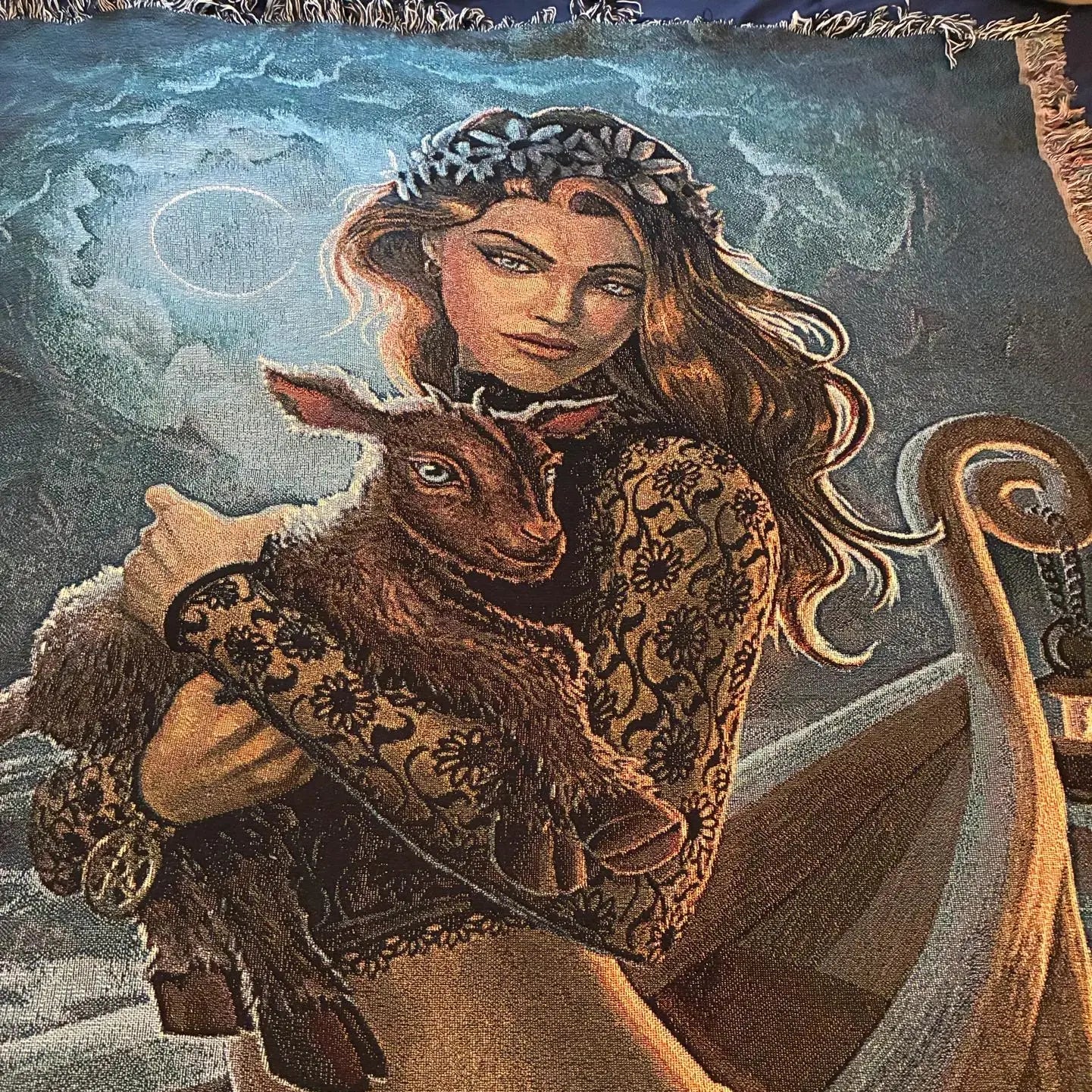 A close-up of a woven tapestry featuring an illustrated scene of a woman in a floral crown holding a brown lamb, with a boat and moonlit sky in the background.