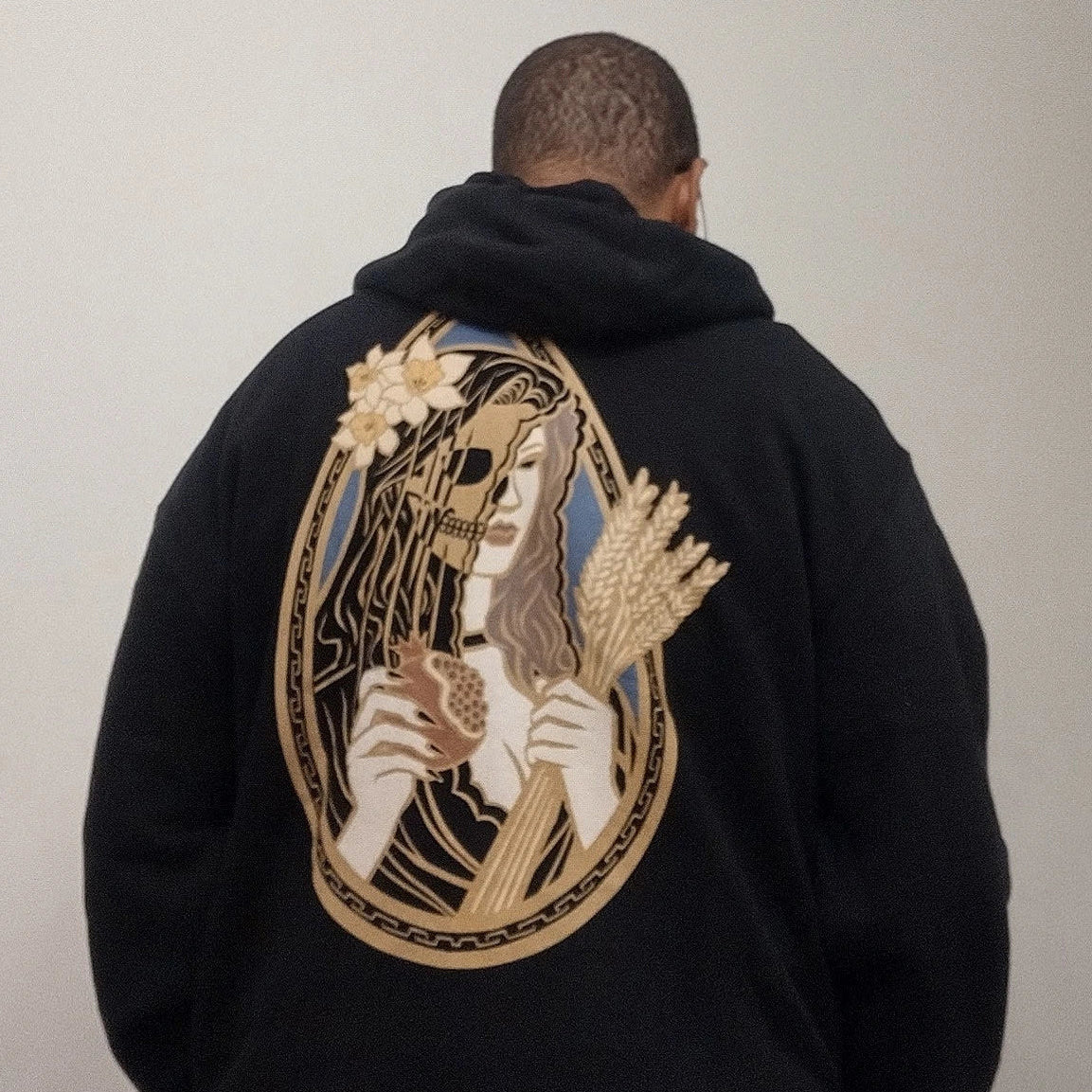 A person viewed from behind wearing a black hoodie with a large graphic on the back featuring a stylized portrait of Persephone in a gold teardrop shaped frame.