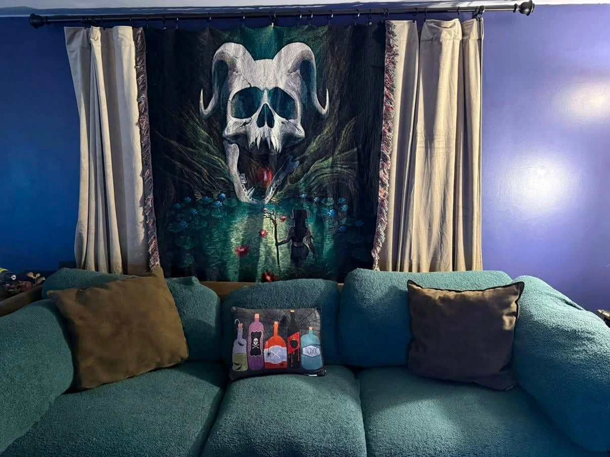 A large wall tapestry featuring a skull with curved horns over a dark forest scene, hung on a window above a teal sofa with decorative pillows.