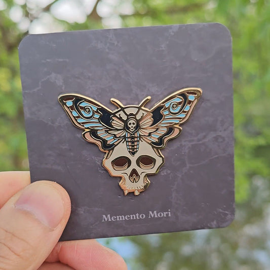 Gold hard enamel pin featuring a death’s head moth resting on a skull