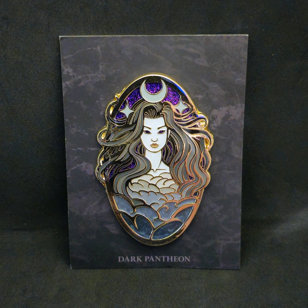 An enamel pin featuring a dark fantasy female figure with long wavy hair, a crescent moon and star crown, and a cloud-like patterned bodice, mounted on a dark gray card labeled 'DARK PANTHEON' against a dark background.