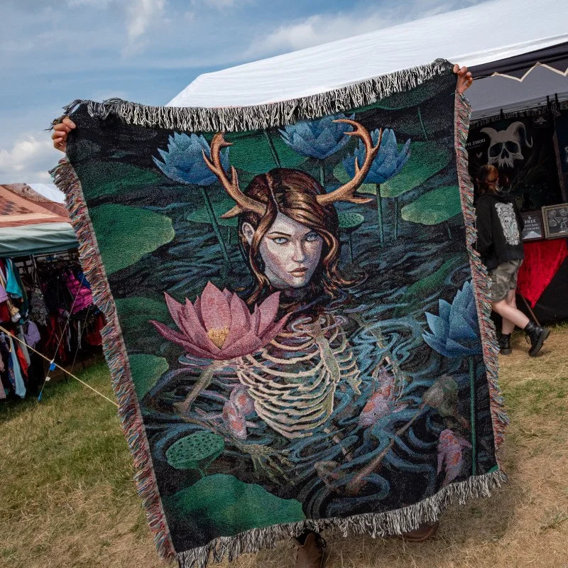 A large woven blanket or throw is held up at an outdoor market. The blanket features a dark fantasy illustration of a skeletal female figure with antlers in a pond, surrounded by blue lotus flowers and green lily pads.
