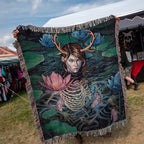 A large woven blanket or throw is held up at an outdoor market. The blanket features a dark fantasy illustration of a skeletal female figure with antlers in a pond, surrounded by blue lotus flowers and green lily pads.